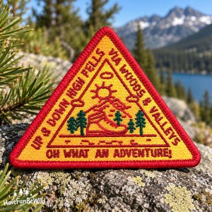 Könnte beinhalten: Ein dreieckiger gestickter Aufnäher mit rotem Rand und gelbem Hintergrund. Er zeigt einen roten Wanderschuh, grüne Kiefern, eine Sonne und den Text: UP & DOWN HIGH FELLS VIA WOODS & VALLEYS OH WHAT AN ADVENTURE! Das Design ist in Rot und Grün gestickt.