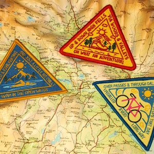 Peut inclure: Trois patchs brodés triangulaires avec des bordures jaunes et des centres bleus. Les patchs représentent différentes activités de plein air : la natation, la randonnée et le vélo. Le texte sur les patchs est : « It was wild & exhilarating and just a little bit chilly I swam in the open water », « Up & down high fells via woods & valleys Oh what an adventure » et « Over passes & through dales I pedalled a very long way A few cake stops that day ».