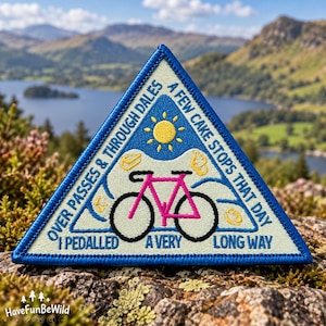 May include: A triangular embroidered patch with a blue border, featuring a pink bicycle, a yellow sun, and small food icons. The text reads: OVER PASSES & THROUGH DALES A FEW CAKE STOPS THAT DAY I PEDALLED A VERY LONG WAY on a cream background.