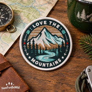 May include: A circular embroidered patch featuring a mountain landscape with a winding river, pine trees, and a sunset sky. The text "LOVE THE MOUNTAINS" is stitched in white along the border. The patch has a dark green background with a white edge.