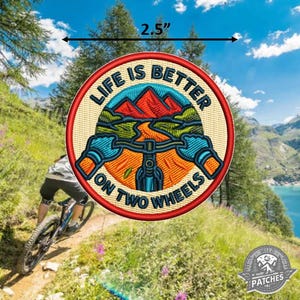 May include: A circular embroidered patch with a red border, featuring a mountain biking scene with orange mountains, green hills, and blue handlebars. The text reads LIFE IS BETTER ON TWO WHEELS. A black arrow indicates a width of 6.35 cm.
