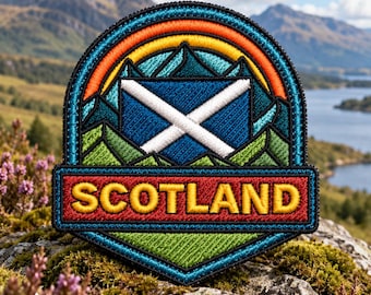 May include: A circular embroidered patch featuring a blue and white Scottish flag centered over stylized green mountains. An orange and yellow arc sits above the mountains. The word SCOTLAND is stitched in yellow letters on a red rectangular base.