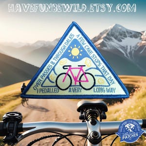 Pode incluir: Um remendo triangular azul e amarelo com uma bicicleta e o texto "Over passes & through dales, a few cake stops that day, I pedalled a very long way". O remendo está no guidão de uma bicicleta de montanha.