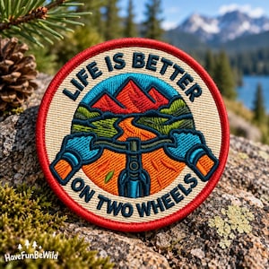 May include: A circular embroidered patch with a red border and a cream background. It features a mountain biking scene with red mountains, green hills, and an orange trail. The text reads LIFE IS BETTER ON TWO WHEELS in dark blue lettering.