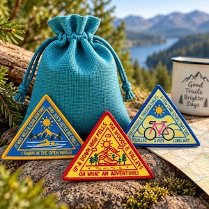 Può includere: Una pochette con coulisse color verde acqua in tessuto testurizzato, accompagnata da tre toppe ricamate triangolari. Le toppe presentano temi all'aperto con testi come 'I swam in the open water', 'Oh what an adventure!' e 'A very long way'.