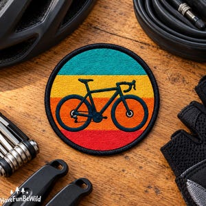 Pode incluir: Um emblema bordado circular com a silhueta de uma bicicleta azul-marinho escuro centrada sobre faixas horizontais em turquesa, amarelo, laranja e vermelho. O emblema tem uma borda preta costurada e destina-se a ser aplicado em tecidos.