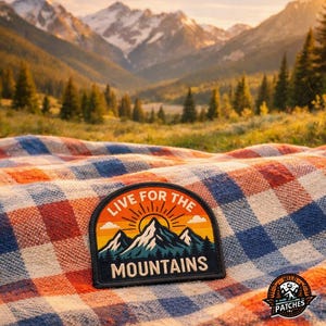 May include: A semi-circular embroidered patch featuring a mountain landscape with a rising sun, orange sky, and dark blue peaks. The text "LIVE FOR THE MOUNTAINS" is stitched in white lettering. The patch is placed on a red, white, and blue plaid fabric.