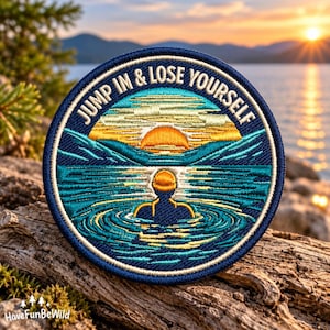 May include: A circular embroidered patch featuring a dark blue border and the text JUMP IN & LOSE YOURSELF. The design depicts a person swimming in a lake towards a bright orange sunset over distant mountains, rendered in shades of blue, teal, and gold.