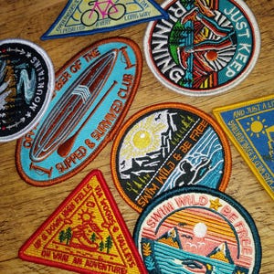 May include: A selection of embroidered patches with outdoor and adventure themes. Designs include a bicycle, mountains, a paddleboard, and phrases like "Swim Wild & Be Free" and "Just Keep Running". The patches are in various shapes and colours.
