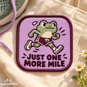 Può includere: Una toppa ricamata quadrata con angoli arrotondati raffigurante una rana verde che corre con pantaloncini viola e scarpe da ginnastica bianche. Lo sfondo è viola chiaro con il testo JUST ONE MORE MILE in lettere viola scuro. La toppa ha un bordo viola scuro.