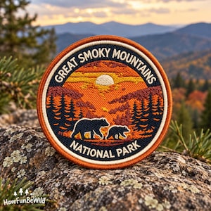 May include: A circular embroidered patch featuring a sunset landscape with a bear and a cub in the foreground. The text GREAT SMOKY MOUNTAINS is arched at the top, and NATIONAL PARK is at the bottom. The design uses orange, yellow, and dark blue.