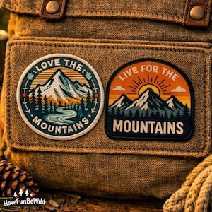 May include: A brown canvas bag featuring two embroidered patches. The circular patch displays a mountain range, a river, and the text LOVE THE MOUNTAINS. The arched patch shows a sun rising over mountains with the text LIVE FOR THE MOUNTAINS.