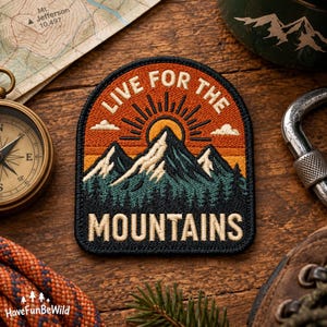 Op de afbeelding: Een geborduurde patch met een zwarte rand met een bergketen in donkergroen en wit, een opkomende zon in oranje en geel, en de tekst "LIVE FOR THE MOUNTAINS" in crèmekleurige letters tegen een oranje luchtachtergrond.