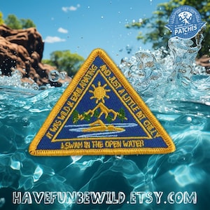 May include: A triangular yellow and blue Girl Scout patch with the text "It was wild & exhilarating and just a little bit chilly. I swam in the open water." The patch is partially submerged in water.