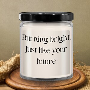 Puede incluir: Una vela en tarro de cristal con tapa negra. La vela tiene una etiqueta blanca con el texto "Burning bright, just like your future" en negro. La vela está sobre un posavasos de madera.