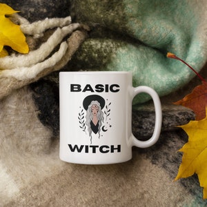May include: White ceramic mug with the words "BASIC WITCH" in black block letters. The mug features a graphic of a woman with long grey hair wearing a black hat. The mug is set against a backdrop of autumn leaves and a cosy blanket.