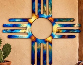 Puede incluir: Una pieza de arte mural de metal con el diseño del símbolo solar Zia. El objeto tiene un centro circular con cuatro conjuntos de tres barras radiantes. La superficie muestra un acabado tratado térmicamente con gradientes de azul, turquesa y oro.