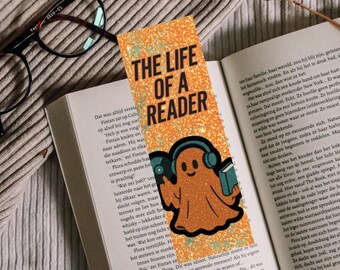 Puede incluir: Un marcapáginas naranja brillante con el texto "THE LIFE OF A READER". Presenta un fantasma de dibujos animados con auriculares y un libro. También se ven unas gafas y un libro abierto.