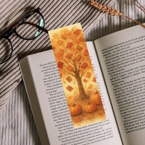 Könnte beinhalten: Herbstliches Lesezeichen mit einem Baum, dessen Blätter Bücher sind, und Kürbissen am Fuß. Das Lesezeichen liegt auf einem offenen Buch. Die Farbpalette umfasst Orange, Braun und Gelb. Das Lesezeichen ist etwa 18 cm lang.