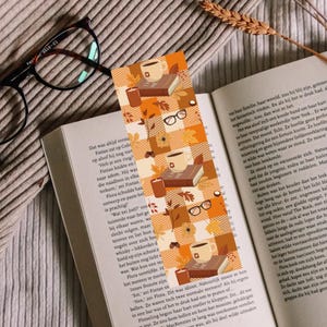May include: A rectangular paper bookmark featuring a repeating pattern of orange and white plaid squares. The design includes illustrations of coffee mugs, stacked books, eyeglasses, candles, and autumn leaves scattered across the surface.
