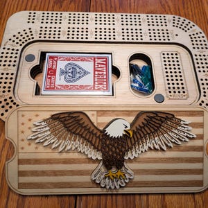 May include: A wooden cribbage board with a detailed eagle design and an American flag motif. The board includes a deck of playing cards and pegs. The word "MAVERICK" is visible on the card box.