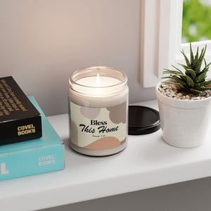 May include: A lit candle in a glass jar with a black lid. The label reads "Bless This Home" with a soft, neutral color palette. A small succulent in a white pot sits nearby on a white shelf.