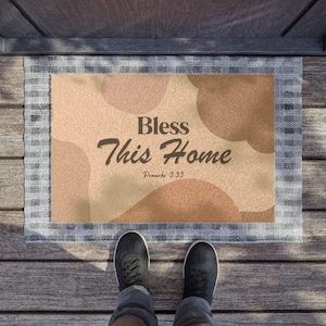May include: A rectangular doormat with the text "Bless This Home" and "Proverbs 3:33" in dark brown script. The mat has a beige background with brown cloud-like shapes and a gray and white checkered border. Black shoes are visible.