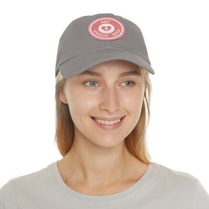Op de afbeelding: Grijze baseballpet met een cirkelvormige patch met de woorden "ASK", "KNOCK" en "SEEK" rond een kruissymbool. De patch heeft een roze rand en een witte achtergrond. De pet wordt door een persoon gedragen.