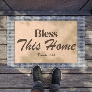 May include: A rectangular doormat with the words "Bless This Home" and the Bible verse Proverbs 3:33 in dark brown script. The mat has a beige center and a blue and white plaid border. Black shoes and blue jeans are visible.