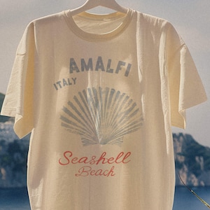 May include: A cream-colored short-sleeve t-shirt featuring a graphic print with the text Amalfi Italy and Seashell Beach in blue and red lettering, centered above a blue illustration of a seashell, hanging on a white hanger against a bright sky.