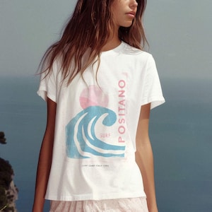 May include: A white short-sleeved t-shirt featuring a graphic print of a blue wave, a pink sun, and the vertical text POSITANO in pink. Below the wave, smaller text reads SURF and SURF CAMP ITALY 1992. The shirt is paired with a white lace skirt.