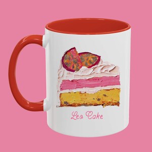 May include: White ceramic mug with a red rim and handle. The mug features a painting of a slice of cake with pink frosting and passion fruit on top. The text "Leo Cake" is printed below the cake.