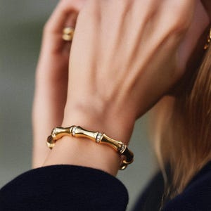 Pode incluir: Uma caneca de cerâmica branca com uma alça preta e o texto "But First, Coffee" em negrito preto. A caneca está sobre um fundo desfocado, sugerindo um ambiente acolhedor.