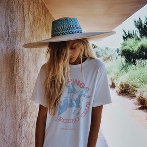 May include: A white t-shirt with a pink and blue graphic of a cowboy riding a horse. The graphic says "Ridin's Rodeo Club 1990 California". The person wearing the shirt is also wearing a white wide-brimmed hat with a blue and white patterned band.