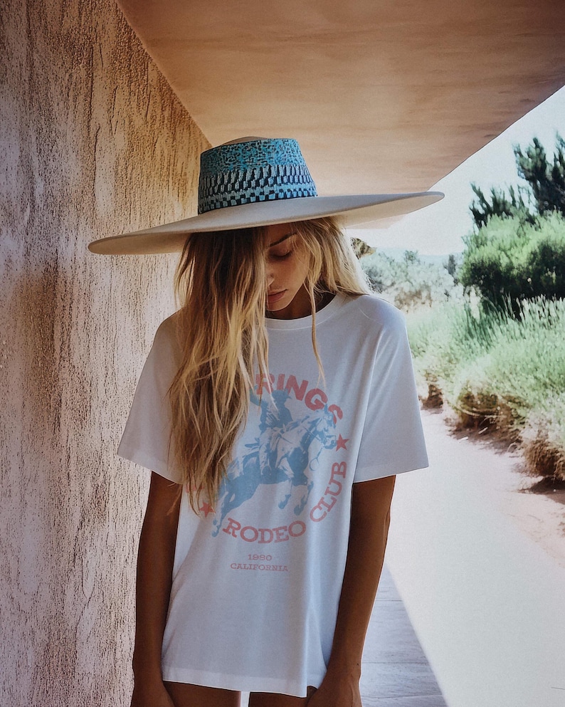May include: A white t-shirt with a pink and blue graphic of a cowboy riding a horse. The graphic says "Ridin's Rodeo Club 1990 California". The person wearing the shirt is also wearing a white wide-brimmed hat with a blue and white patterned band.