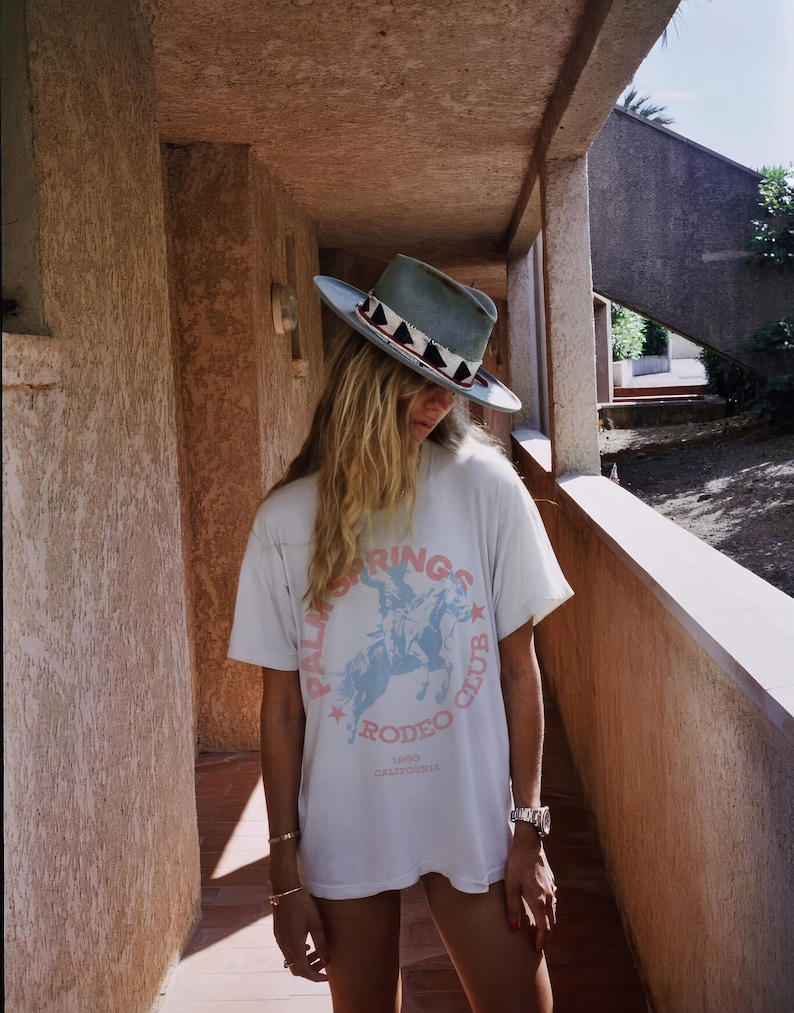 May include: A woman wearing a white t-shirt with a blue and pink graphic of a person riding a horse. The graphic says "Palm Springs Rodeo Club 1980 California". She is wearing a light blue cowboy hat with a black and white patterned band.