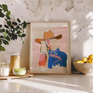 May include: A framed art print featuring an abstract illustration of a figure wearing a tan cowboy hat. The artwork uses a colour palette of soft pink, vibrant blue, and orange brushstrokes against a light background, presented in a light wood frame.