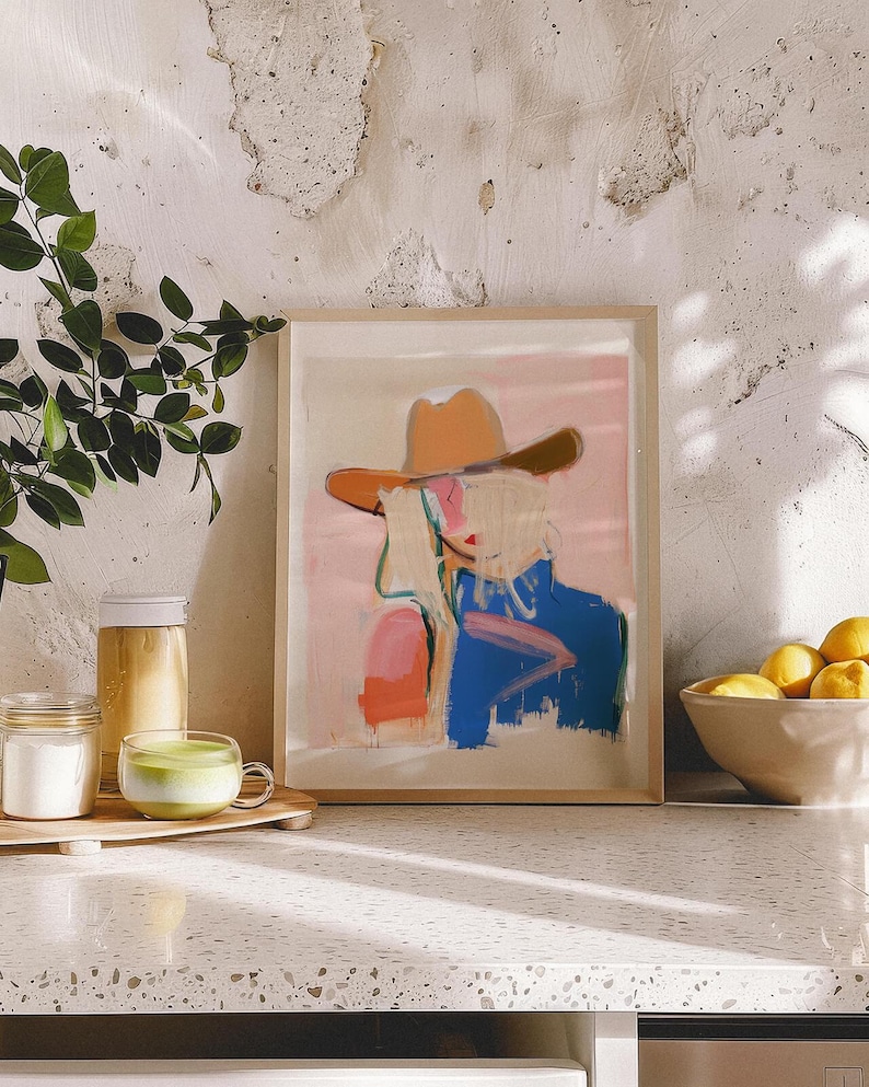 May include: A framed art print featuring an abstract illustration of a figure wearing a tan cowboy hat. The artwork uses a colour palette of soft pink, vibrant blue, and orange brushstrokes against a light background, presented in a light wood frame.