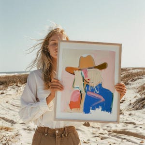 May include: A framed art print featuring an abstract painting of a figure wearing a tan cowboy hat. The artwork uses a palette of soft pink, bright blue, and orange brushstrokes against a light background, encased in a light wood frame.