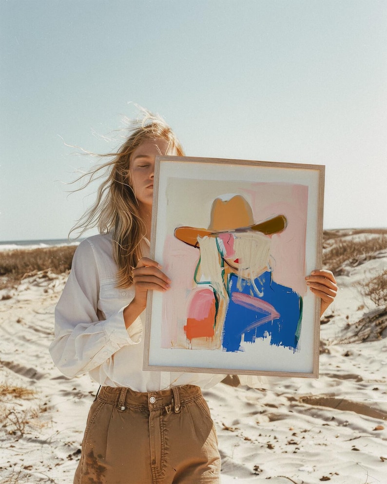 May include: A framed art print featuring an abstract painting of a figure wearing a tan cowboy hat. The artwork uses a palette of soft pink, bright blue, and orange brushstrokes against a light background, encased in a light wood frame.