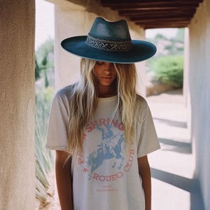 May include: A woman wearing a white t-shirt with a blue and pink graphic that says "Palm Springs Rodeo Club 1950 California". She is wearing a blue wide-brimmed hat with a woven band.
