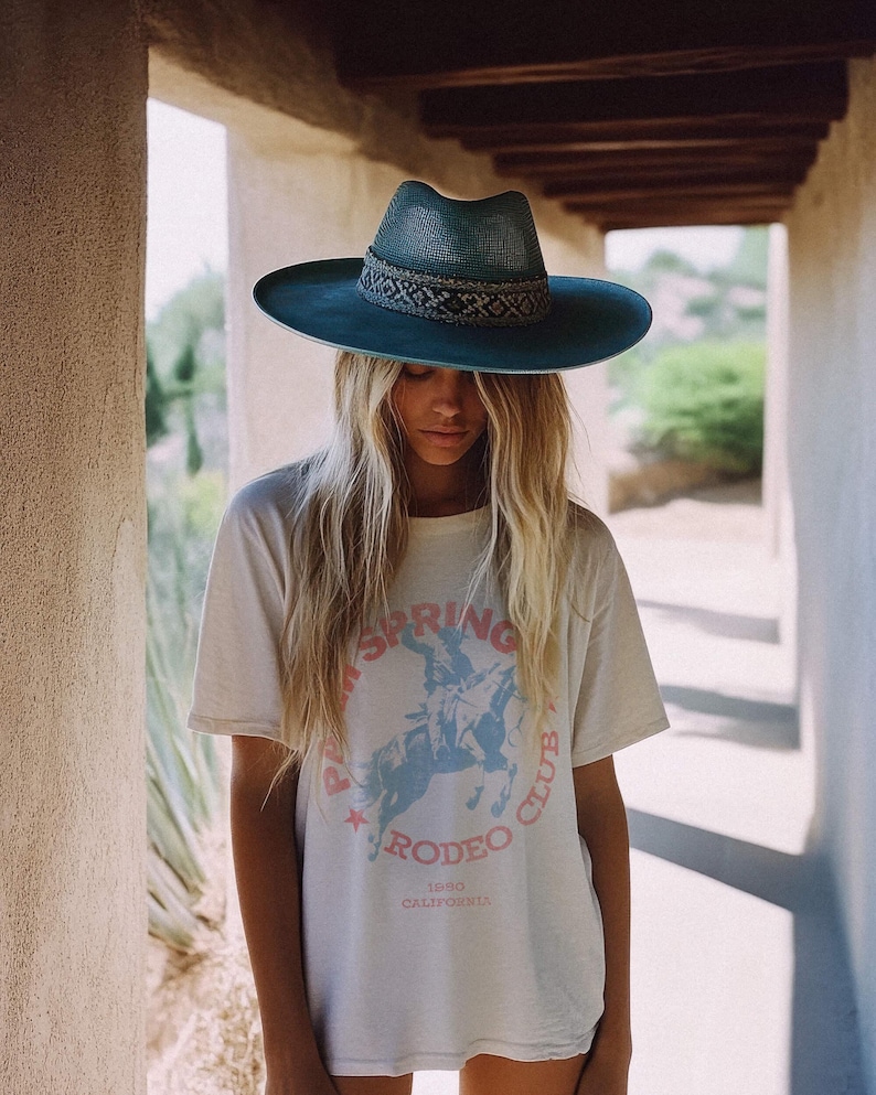 May include: A woman wearing a white t-shirt with a blue and pink graphic that says "Palm Springs Rodeo Club 1950 California". She is wearing a blue wide-brimmed hat with a woven band.
