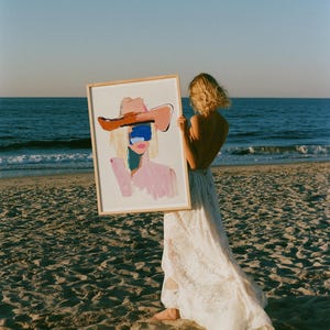 Puede incluir: Una impresi&oacute;n art&iacute;stica rectangular enmarcada que presenta una pintura abstracta de una figura con un sombrero de ala ancha. La obra utiliza pinceladas audaces en tonos rosa, marr&oacute;n y un bloque azul sobre los ojos, sobre fondo blanco.