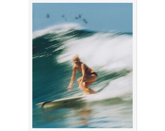 Può includere: Fotografia sfocata di un surfista che cavalca un'onda. Il surfista, in bikini nero, è in equilibrio su una tavola da surf. L'onda è un mix di schiuma bianca e acqua turchese, su uno sfondo di cielo azzurro.