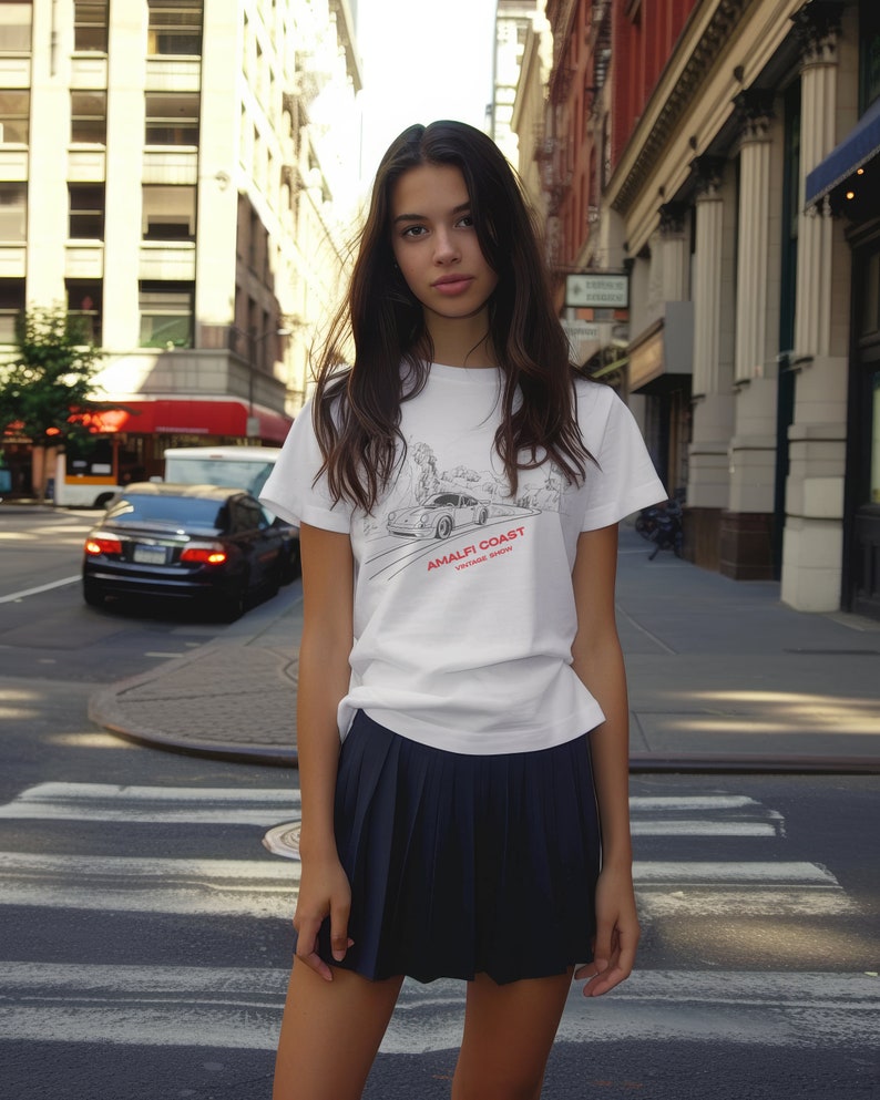 May include: A white t-shirt with a black and white illustration of a car and the text "Amalfi Coast Vintage Show". The person wearing the shirt is standing on a city street.