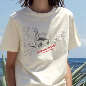 May include: White t-shirt with a black and white illustration of a car driving on a winding road with the text "Amalfi Coast Vintage Show" printed on the front.