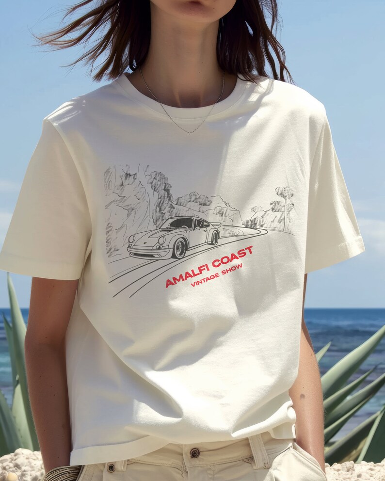 May include: White t-shirt with a black and white illustration of a car driving on a winding road with the text "Amalfi Coast Vintage Show" printed on the front.