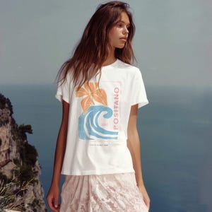 May include: A white short-sleeve t-shirt featuring a graphic print of an orange flower and a blue ocean wave. The text POSITANO is printed vertically in pink, with the words SURF and ITALY CAMP 1990 printed in smaller black lettering.