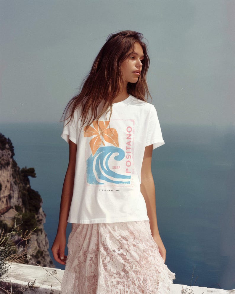 May include: A white short-sleeve t-shirt featuring a graphic print of an orange flower and a blue ocean wave. The text POSITANO is printed vertically in pink, with the words SURF and ITALY CAMP 1990 printed in smaller black lettering.
