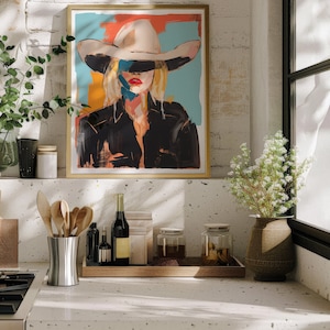 May include: A framed painting of a woman wearing a white cowboy hat and a black leather jacket. The background is a colorful abstract design. The painting is hanging on a white brick wall above a kitchen counter.